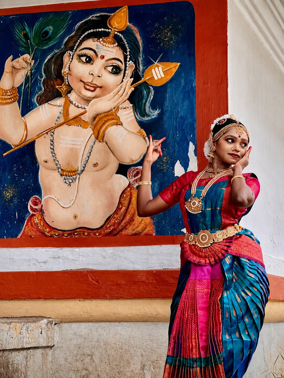 Lakshmi Kumaran in Bharatanatyam costume posing before a colourful temple mural